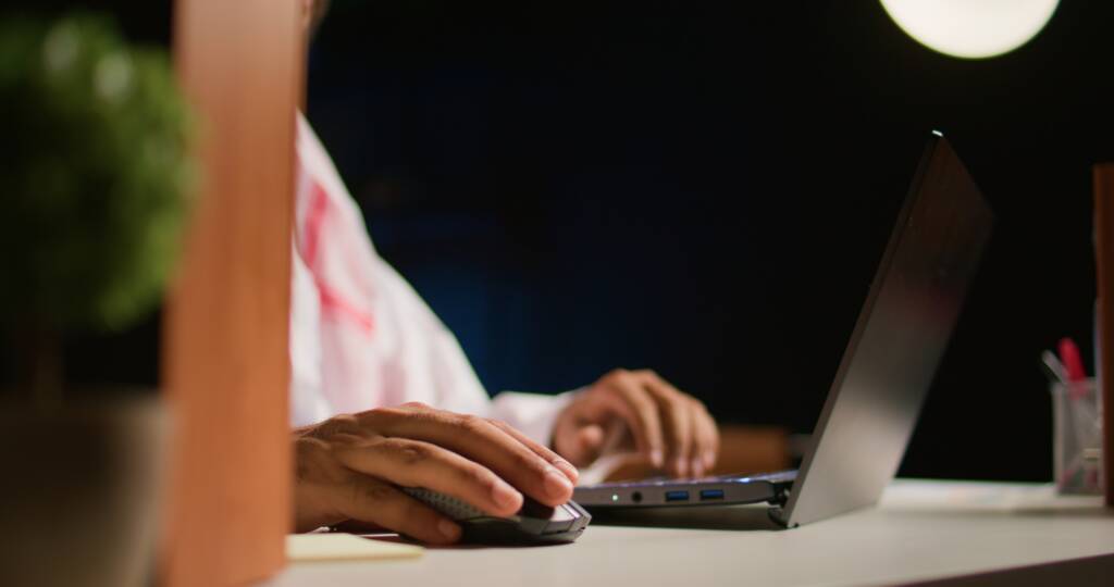 Person arbeitet nachts am Laptop, eine Hand an der Maus, Schreibtischlampe als einzige Lichtquelle